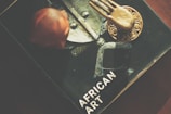 A book titled 'African Art' is partially visible, featuring a central image of a metallic and textured object. On top of the book, there is a bronze or gold-colored decorative item with a hand-like design, and a reddish-brown carved figure.