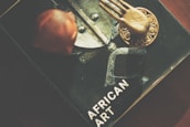 A book titled 'African Art' is partially visible, featuring a central image of a metallic and textured object. On top of the book, there is a bronze or gold-colored decorative item with a hand-like design, and a reddish-brown carved figure.