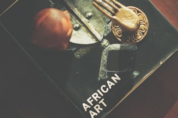 A book titled 'African Art' is partially visible, featuring a central image of a metallic and textured object. On top of the book, there is a bronze or gold-colored decorative item with a hand-like design, and a reddish-brown carved figure.