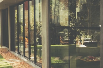 A modern building with large glass windows reflects a lush outdoor scene, featuring trees and greenery. Inside, there are hints of a cozy interior with visible furnishings, such as chairs and a table. The outdoor area is landscaped with rocks and grass, creating a harmonious blend of nature and architecture.