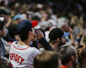 A person wearing a baseball jersey with the name 'Spidey' and a cap is seen in a crowd of people at a sporting event. The individual appears to be cheering or shouting enthusiastically. The scene is lively with many spectators visible, wearing casual attire such as hats and t-shirts.
