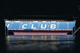 Stadium seating is visible from the perspective of a tunnel entrance. The seats form the word 'CLUB' in large, white letters against a backdrop of blue seats. The atmosphere suggests an anticipation of entering the main stadium area.