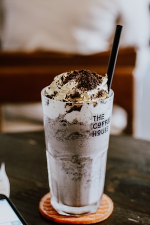 A frosty beverage with a generous topping of whipped cream and chocolate sprinkles is served in a tall glass with a black straw. The glass has 'The Coffee House' printed on it and rests on a round coaster atop a dark wooden table. The background features a blurred indoor setting, creating a cozy ambiance.
