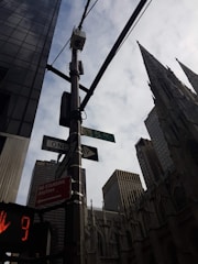 A cityscape featuring tall buildings and a prominent architectural structure with a gothic design. The scene includes street signs indicating Fifth Avenue and a one-way street, along with a traffic signal displaying a red hand with a countdown. Overhead, wires and a security camera are attached to a pole, and the sky is partly cloudy.