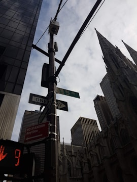 A cityscape featuring tall buildings and a prominent architectural structure with a gothic design. The scene includes street signs indicating Fifth Avenue and a one-way street, along with a traffic signal displaying a red hand with a countdown. Overhead, wires and a security camera are attached to a pole, and the sky is partly cloudy.