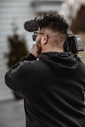 A person holding and filming with a professional video camera, wearing a black hoodie, and standing outside with blurred background elements.