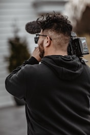 A person holding and filming with a professional video camera, wearing a black hoodie, and standing outside with blurred background elements.