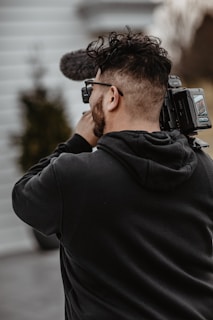 A person holding and filming with a professional video camera, wearing a black hoodie, and standing outside with blurred background elements.