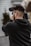 A person holding and filming with a professional video camera, wearing a black hoodie, and standing outside with blurred background elements.