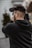 A person holding and filming with a professional video camera, wearing a black hoodie, and standing outside with blurred background elements.