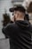 A person holding and filming with a professional video camera, wearing a black hoodie, and standing outside with blurred background elements.