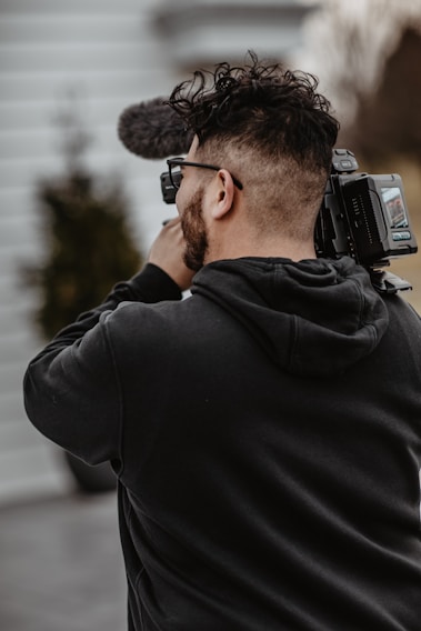 A person holding and filming with a professional video camera, wearing a black hoodie, and standing outside with blurred background elements.