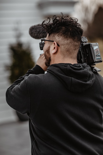 A person holding and filming with a professional video camera, wearing a black hoodie, and standing outside with blurred background elements.