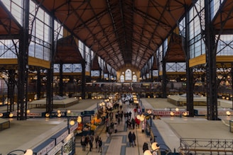 An indoor market hall with a high, arched ceiling supported by steel beams, creating an industrial yet ornate architectural style. The hall is bustling with people walking along a central aisle lined with various kiosks and market stalls. Some stalls are selling fruits and vegetables, with vibrant displays of produce. Overhead lights illuminate the space, adding a warm glow to the bustling environment.