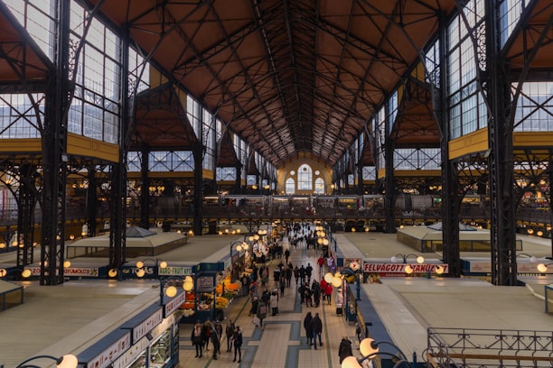 An indoor market hall with a high, arched ceiling supported by steel beams, creating an industrial yet ornate architectural style. The hall is bustling with people walking along a central aisle lined with various kiosks and market stalls. Some stalls are selling fruits and vegetables, with vibrant displays of produce. Overhead lights illuminate the space, adding a warm glow to the bustling environment.