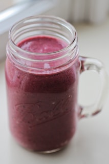 A clear mason jar filled with a thick, vibrant purple smoothie. The jar has a sturdy handle and a slightly embossed logo. The smoothie looks smooth and creamy, suggesting it's blended well. Light from a nearby window softly illuminates the jar, casting a gentle shadow on the surface.