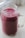 A clear mason jar filled with a thick, vibrant purple smoothie. The jar has a sturdy handle and a slightly embossed logo. The smoothie looks smooth and creamy, suggesting it's blended well. Light from a nearby window softly illuminates the jar, casting a gentle shadow on the surface.
