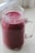 A clear mason jar filled with a thick, vibrant purple smoothie. The jar has a sturdy handle and a slightly embossed logo. The smoothie looks smooth and creamy, suggesting it's blended well. Light from a nearby window softly illuminates the jar, casting a gentle shadow on the surface.