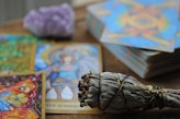 A collection of colorful tarot cards is spread out on a wooden surface, featuring intricate artwork and vibrant imagery. A bundle of sage wrapped with string is placed in the foreground, while a blurred crystal sits behind the cards.