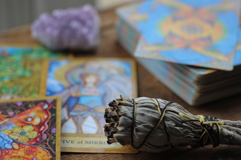 A collection of colorful tarot cards is spread out on a wooden surface, featuring intricate artwork and vibrant imagery. A bundle of sage wrapped with string is placed in the foreground, while a blurred crystal sits behind the cards.