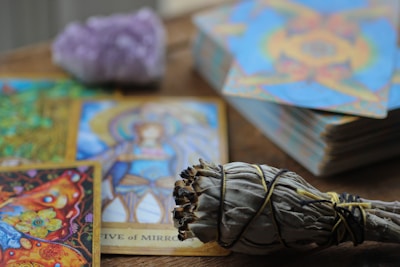 A collection of colorful tarot cards is spread out on a wooden surface, featuring intricate artwork and vibrant imagery. A bundle of sage wrapped with string is placed in the foreground, while a blurred crystal sits behind the cards.