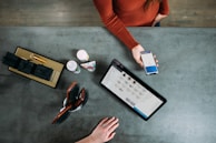 A person is holding a smartphone displaying a payment screen over a countertop. A tablet with a product listing is also present, alongside grooming tools, a few bottles, and a box with black-wrapped items. Another hand is resting on the counter.