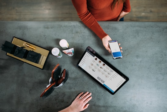 A person is holding a smartphone displaying a payment screen over a countertop. A tablet with a product listing is also present, alongside grooming tools, a few bottles, and a box with black-wrapped items. Another hand is resting on the counter.