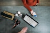 A person is holding a smartphone displaying a payment screen over a countertop. A tablet with a product listing is also present, alongside grooming tools, a few bottles, and a box with black-wrapped items. Another hand is resting on the counter.