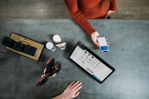 A person is holding a smartphone displaying a payment screen over a countertop. A tablet with a product listing is also present, alongside grooming tools, a few bottles, and a box with black-wrapped items. Another hand is resting on the counter.