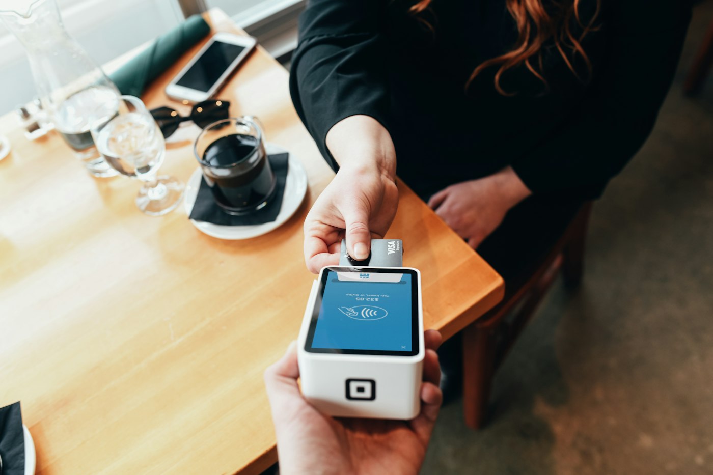 A customer holds a bank card above a handheld Square payment terminal beside a cafe table
