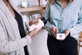 Two people are engaged in a transaction, one holding a small container with a digital payment logo and the other holding a bottle of tea. They are both dressed in casual clothing, standing inside a shop with shelves in the background displaying various products.