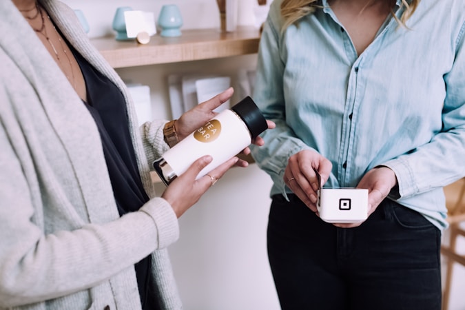 Two people are engaged in a transaction, one holding a small container with a digital payment logo and the other holding a bottle of tea. They are both dressed in casual clothing, standing inside a shop with shelves in the background displaying various products.