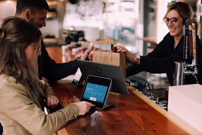 A woman is making a purchase using a card reader at a bar or cafe counter. Another person is handing over a shopping bag to her. The counter has digital payment equipment and beverage taps.