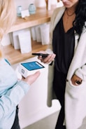 Two people are engaged in a transaction using mobile devices. One person is holding a smartphone, while the other holds a modern card reader with a digital display. They are in a setting that looks like a store or a boutique with shelves in the background containing items and simple decor.