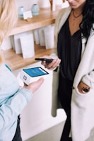 Two people are engaged in a transaction using mobile devices. One person is holding a smartphone, while the other holds a modern card reader with a digital display. They are in a setting that looks like a store or a boutique with shelves in the background containing items and simple decor.