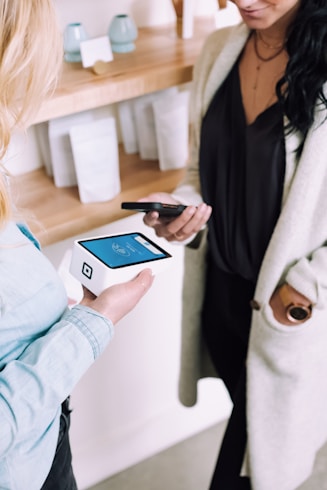Two people are engaged in a transaction using mobile devices. One person is holding a smartphone, while the other holds a modern card reader with a digital display. They are in a setting that looks like a store or a boutique with shelves in the background containing items and simple decor.