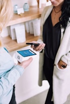 Two people are engaged in a transaction using mobile devices. One person is holding a smartphone, while the other holds a modern card reader with a digital display. They are in a setting that looks like a store or a boutique with shelves in the background containing items and simple decor.