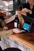 A person is engaged in a transaction at a wooden counter, using a point-of-sale terminal. There is a bottle of wine on the counter and another person is handing over a paper bag, likely containing the purchased item.