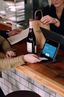 A person is engaged in a transaction at a wooden counter, using a point-of-sale terminal. There is a bottle of wine on the counter and another person is handing over a paper bag, likely containing the purchased item.