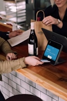 A person is engaged in a transaction at a wooden counter, using a point-of-sale terminal. There is a bottle of wine on the counter and another person is handing over a paper bag, likely containing the purchased item.