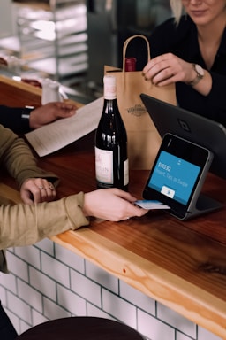 A person is engaged in a transaction at a wooden counter, using a point-of-sale terminal. There is a bottle of wine on the counter and another person is handing over a paper bag, likely containing the purchased item.
