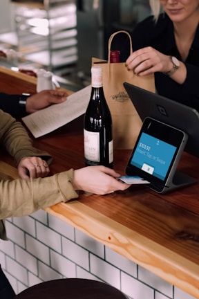 A person is engaged in a transaction at a wooden counter, using a point-of-sale terminal. There is a bottle of wine on the counter and another person is handing over a paper bag, likely containing the purchased item.
