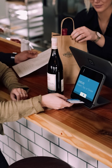 A person is engaged in a transaction at a wooden counter, using a point-of-sale terminal. There is a bottle of wine on the counter and another person is handing over a paper bag, likely containing the purchased item.