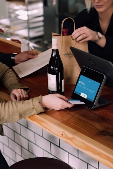 A person is engaged in a transaction at a wooden counter, using a point-of-sale terminal. There is a bottle of wine on the counter and another person is handing over a paper bag, likely containing the purchased item.
