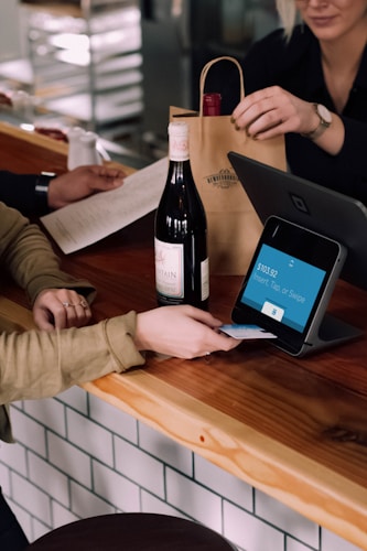 A person is engaged in a transaction at a wooden counter, using a point-of-sale terminal. There is a bottle of wine on the counter and another person is handing over a paper bag, likely containing the purchased item.