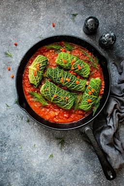 A cast iron skillet filled with a rich tomato sauce, topped with several stuffed cabbage rolls. The rolls are garnished with small pieces of red pepper and sprigs of dill, providing a vibrant contrast to the green cabbage. The skillet is set on a textured dark surface alongside two salt and pepper shakers and a gray cloth napkin.