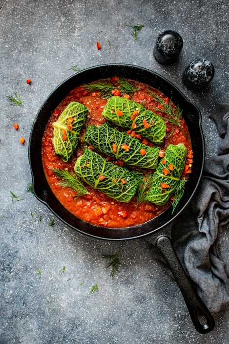 A cast iron skillet filled with a rich tomato sauce, topped with several stuffed cabbage rolls. The rolls are garnished with small pieces of red pepper and sprigs of dill, providing a vibrant contrast to the green cabbage. The skillet is set on a textured dark surface alongside two salt and pepper shakers and a gray cloth napkin.