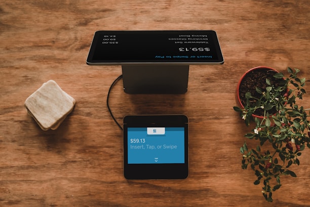 A wooden table holds a black digital payment terminal with a blue screen displaying payment details. Nearby is a potted plant with green leaves on the right, and a rounded square stone object on the left.