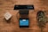 A wooden table holds a black digital payment terminal with a blue screen displaying payment details. Nearby is a potted plant with green leaves on the right, and a rounded square stone object on the left.