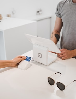 A person is making a payment using a credit card on a small, square card reader connected to a modern point-of-sale system at a minimalist counter. Another person is standing nearby, possibly a cashier, wearing a grey t-shirt. Sunglasses rest on the counter, and the background features a simple, white decor.